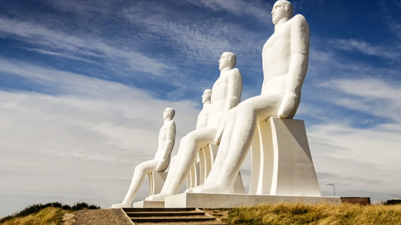 Fire store hvide statuer af siddende menneskefigurer er placeret på en bakke under en delvis overskyet himmel med en jordsti og trapper, der fører op til skulpturerne.