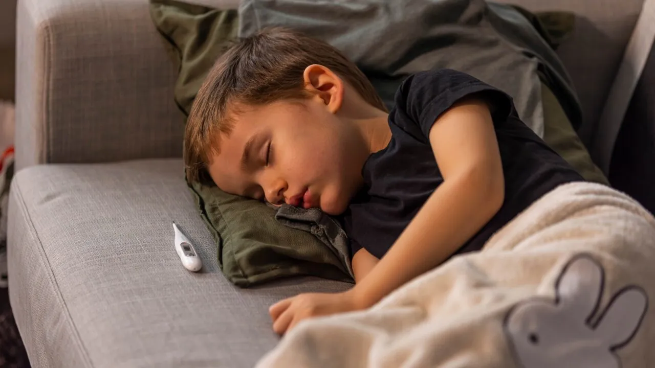 En ung dreng ligger på en sofa og sover med hovedet på en grøn pude. Han er dækket af et tæppe med et kanindesign, og et digitalt termometer ligger ved siden af ham.