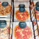 Kasser med frosne pizzaer af mærket Solling, herunder salami-, kebab-, bolognese- og specialevarianter, er stablet i en supermarkedsfryser bag et sort metalstativ.