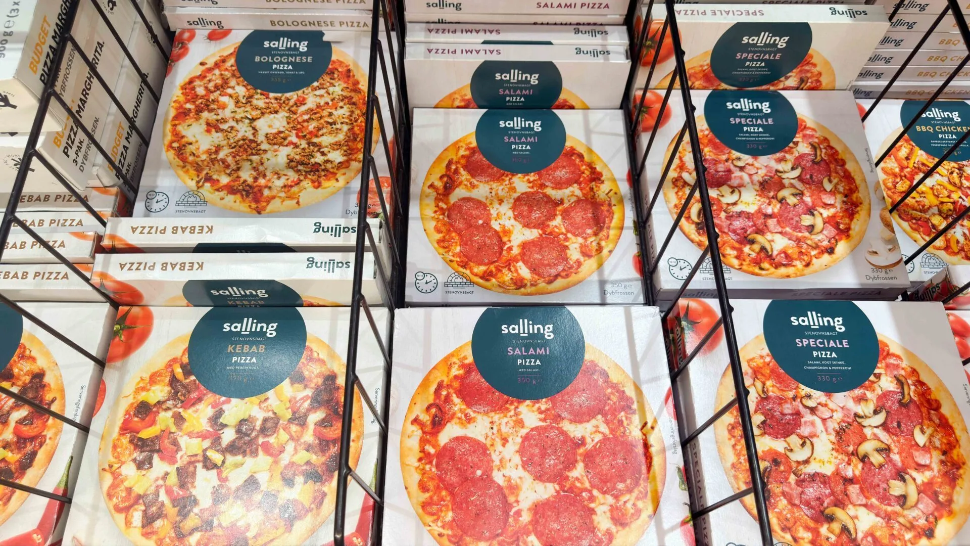 Kasser med frosne pizzaer af mærket Solling, herunder salami-, kebab-, bolognese- og specialevarianter, er stablet i en supermarkedsfryser bag et sort metalstativ.