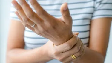 En person iført en stribet skjorte holder sit venstre håndled med højre hånd og ser ud til at have smerter eller ubehag. Personen bærer to guldringe på sin højre hånd.