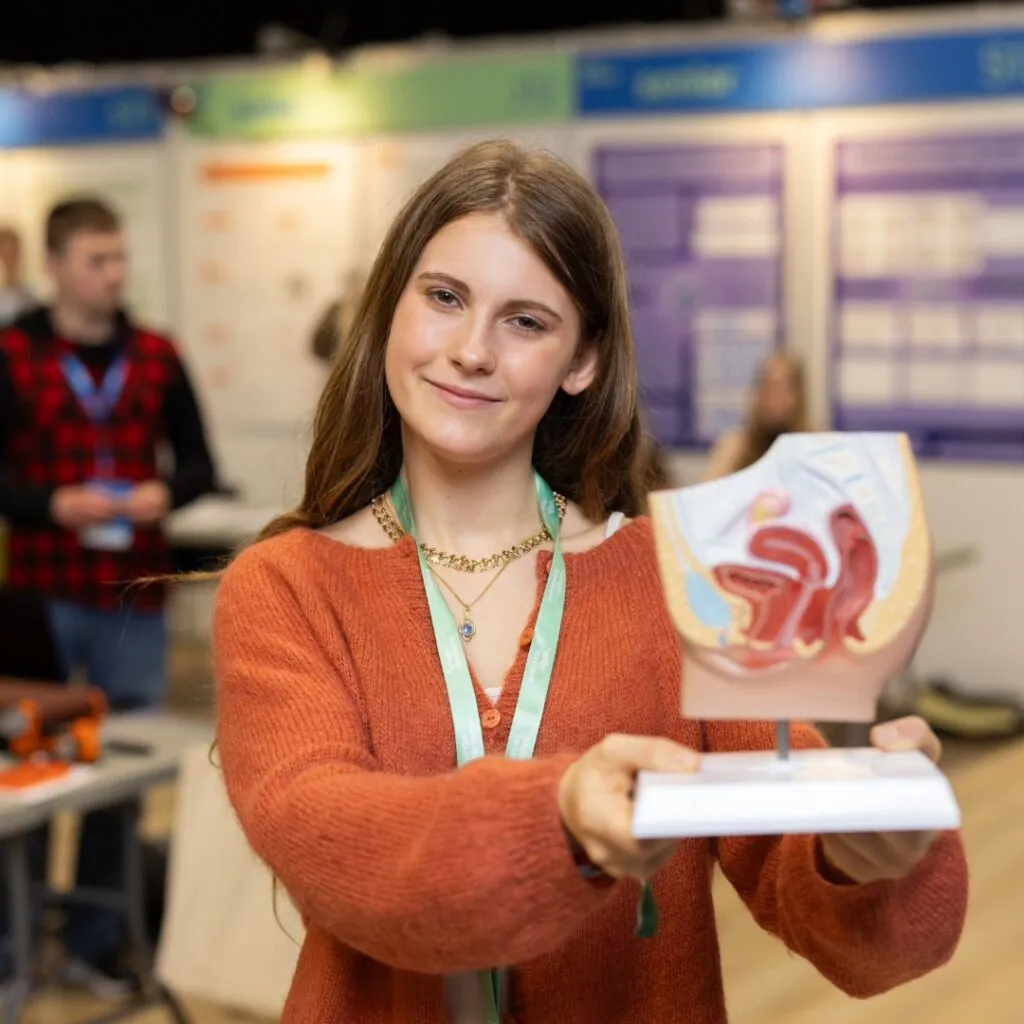 En ung kvinde med langt brunt hår iført en orange sweater smiler og holder en medicinsk model af det kvindelige forplantningssystem op ved en videnskabsmesse eller et uddannelsesarrangement med udstillingstavler i baggrunden.