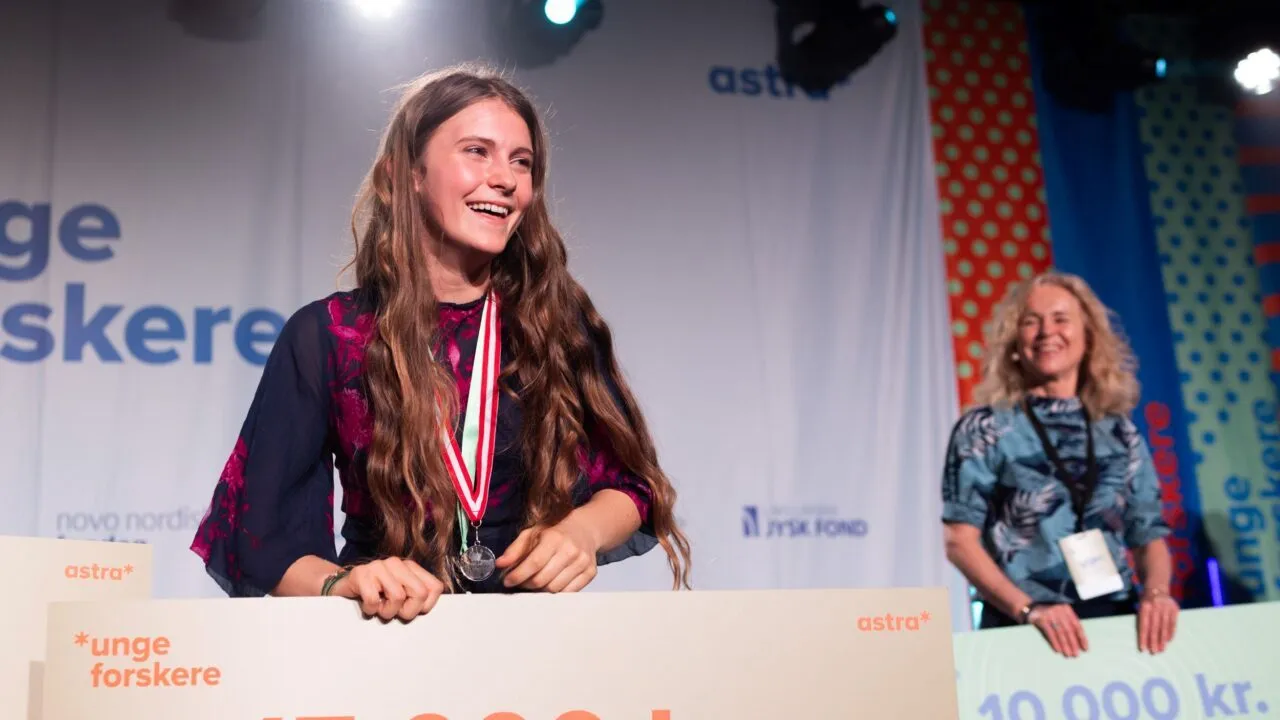 En ung kvinde smiler på scenen med en medalje om halsen og en stor check på 15.000 kr. i hånden. En anden kvinde står i baggrunden og holder en check på 10.000 kr. Bag dem hænger event-bannere.