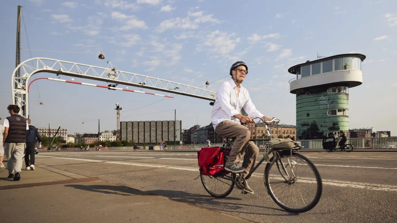 En ældre mand med hjelm og hvid skjorte kører på en cykel med en rød cykeltaske over en bro. I baggrunden ses et grønt, cylindrisk tårn og en delvis overskyet himmel. Andre mennesker går i nærheden.