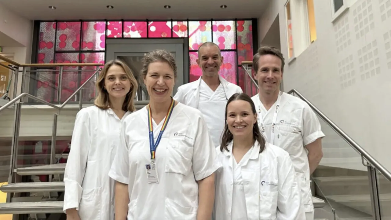 Fem smilende læger i hvide kitler står sammen indendørs foran en trappe og et vindue med et lyserødt, mønstret design.