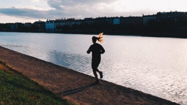 Silhuet af en person, der jogger langs en sti ved en rolig flod i solnedgang, med bygninger og træer synlige på den modsatte bred under en overskyet himmel.