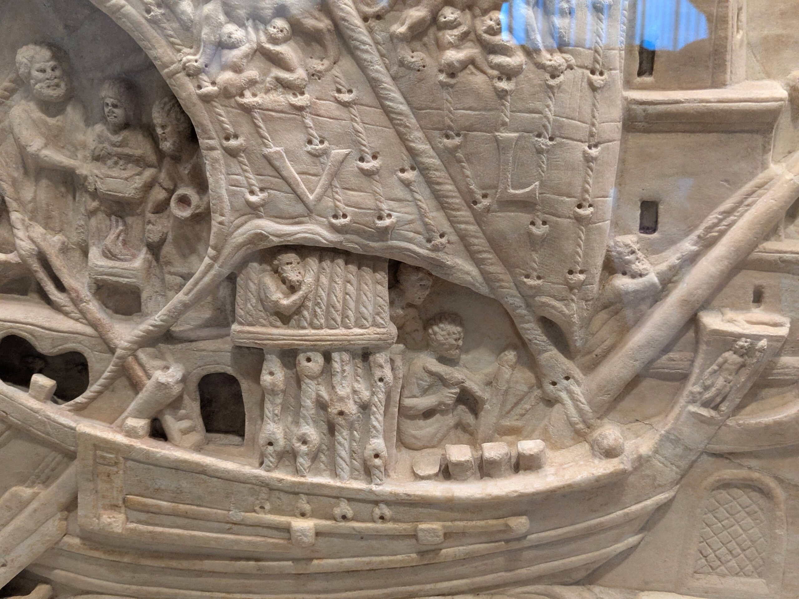 Detaljeret stenskulptur af et gammelt skib med figurer om bord, sejl med V og L, rigning og arkitektoniske detaljer; nogle figurer ser ud til at klatre, arbejde eller interagere.