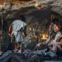 Fire forhistoriske mennesker er inde i en svagt oplyst hule. En person sidder i forgrunden ved et lille bål, mens tre andre står eller bevæger sig i baggrunden. Primitive redskaber og dyrehuder er synlige.