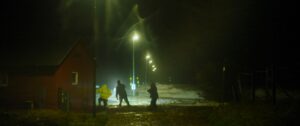 Tre personer står på en våd, mudret vej om natten i nærheden af en rød bygning, der oplyses af en række gadelygter i kraftig tåge eller regn. Stemningen er mørk og tåget med begrænset udsyn.