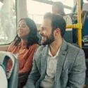 En mand i en grå blazer og en kvinde i en pink skjorte sidder ved siden af hinanden og smiler og taler i en offentlig bus. Andre passagerer sidder i nærheden, og sollyset skinner ind gennem vinduerne.