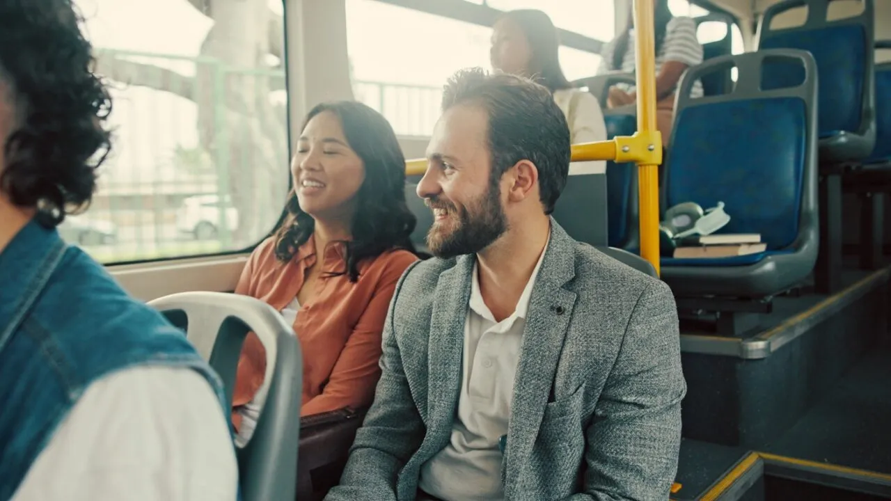 En mand i en grå blazer og en kvinde i en pink skjorte sidder ved siden af hinanden og smiler og taler i en offentlig bus. Andre passagerer sidder i nærheden, og sollyset skinner ind gennem vinduerne.