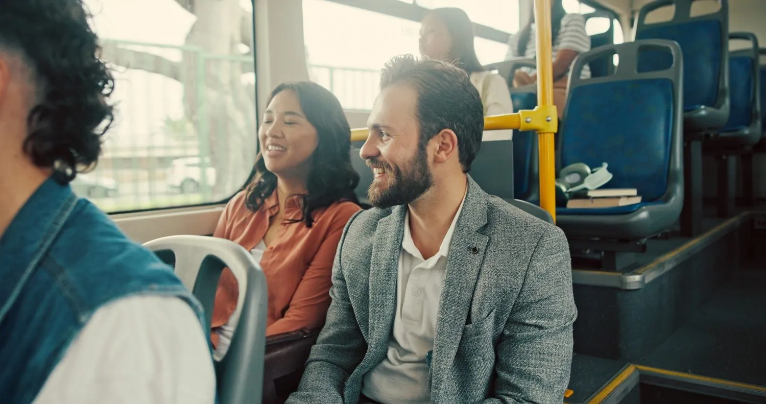 En mand i en grå blazer og en kvinde i en pink skjorte sidder ved siden af hinanden og smiler og taler i en offentlig bus. Andre passagerer sidder i nærheden, og sollyset skinner ind gennem vinduerne.