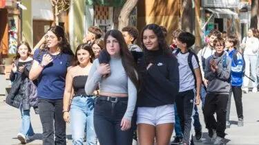 En gruppe teenagere går sammen udendørs på en solskinsdag, nogle taler og smiler. To piger går foran, den ene i grå sweater og jeans, den anden i sort sweatshirt og hvide shorts. Der er butikker i baggrunden.