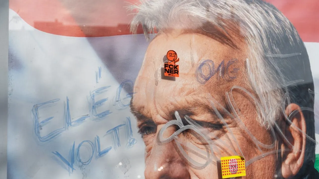 Nærbillede af en mands ansigt på en plakat, der er overmalet med graffiti og to klistermærker. Det ene klistermærke har et orange tegnefilmsansigt og tekst; det andet er gult med røde markeringer. I blå graffiti står der Elég volt! på tværs af billedet.