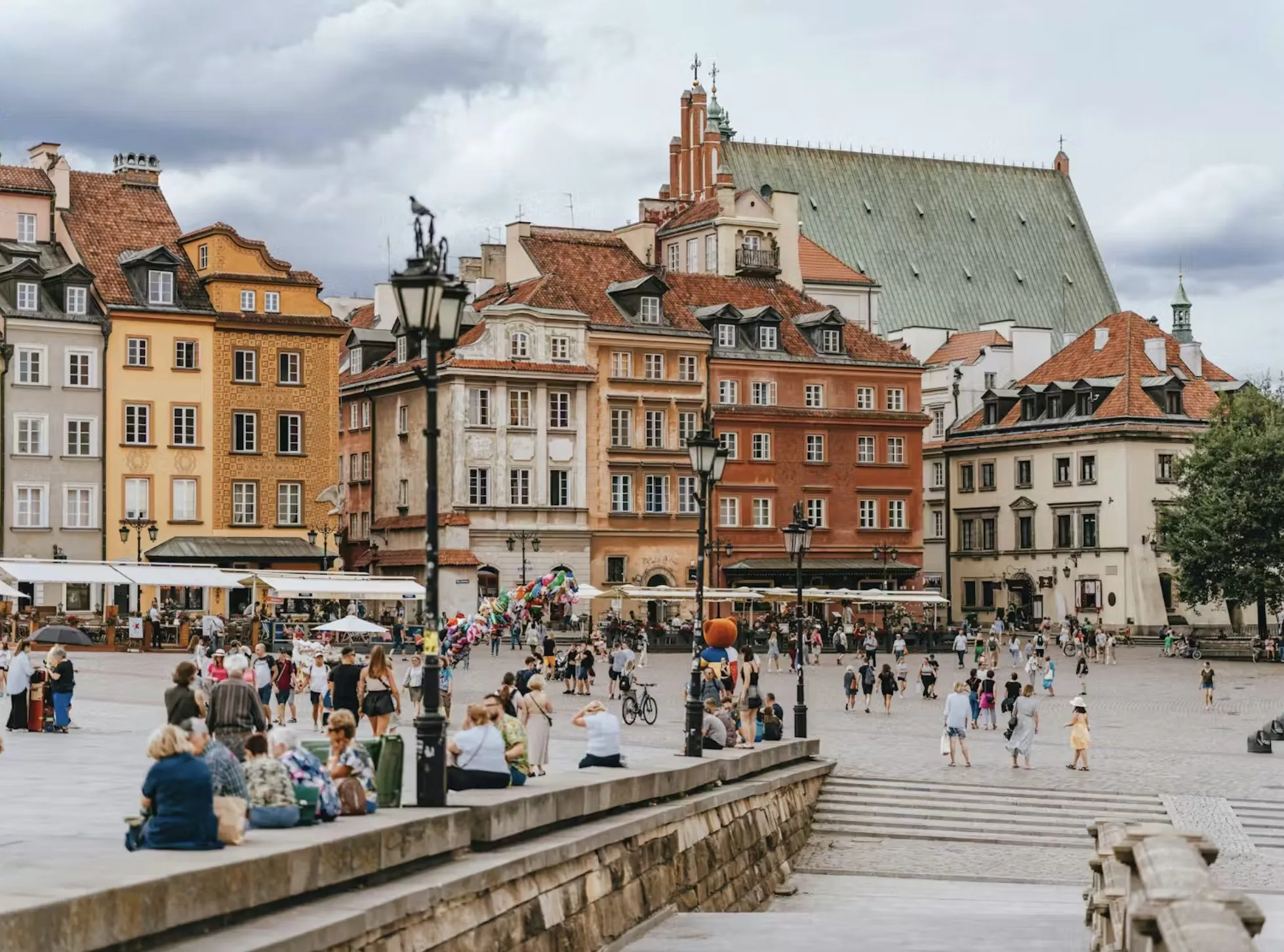 Folk samles på et travlt europæisk torv med farverige historiske bygninger, udendørs caféer og en kirke i baggrunden under en overskyet himmel. Nogle mennesker sidder på trapper, mens andre går eller slapper af på pladsen.