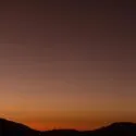 En mørk bjergsilhuet under en grå himmel i skumringen, med dyb orange nær horisonten, der falmer til mørk lilla over. To lyse himmellegemer er synlige tæt på hinanden på himlen.