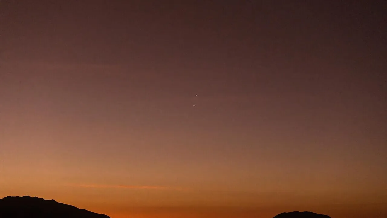 En mørk bjergsilhuet under en grå himmel i skumringen, med dyb orange nær horisonten, der falmer til mørk lilla over. To lyse himmellegemer er synlige tæt på hinanden på himlen.