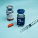 To hætteglas mærket "B12 Vitamin", en orange kapsel og en sprøjte er anbragt på en lyseblå overflade. Hætteglassene har forskelligt farvede låg, et sølvfarvet og et blåt.