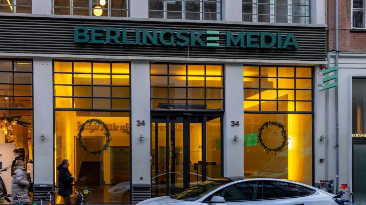 Indgangen til Berlingske Media med store glasdøre og vinduer, der lyser op i et varmt, gult lys. En bil er parkeret foran, og et par mennesker går forbi på fortovet.