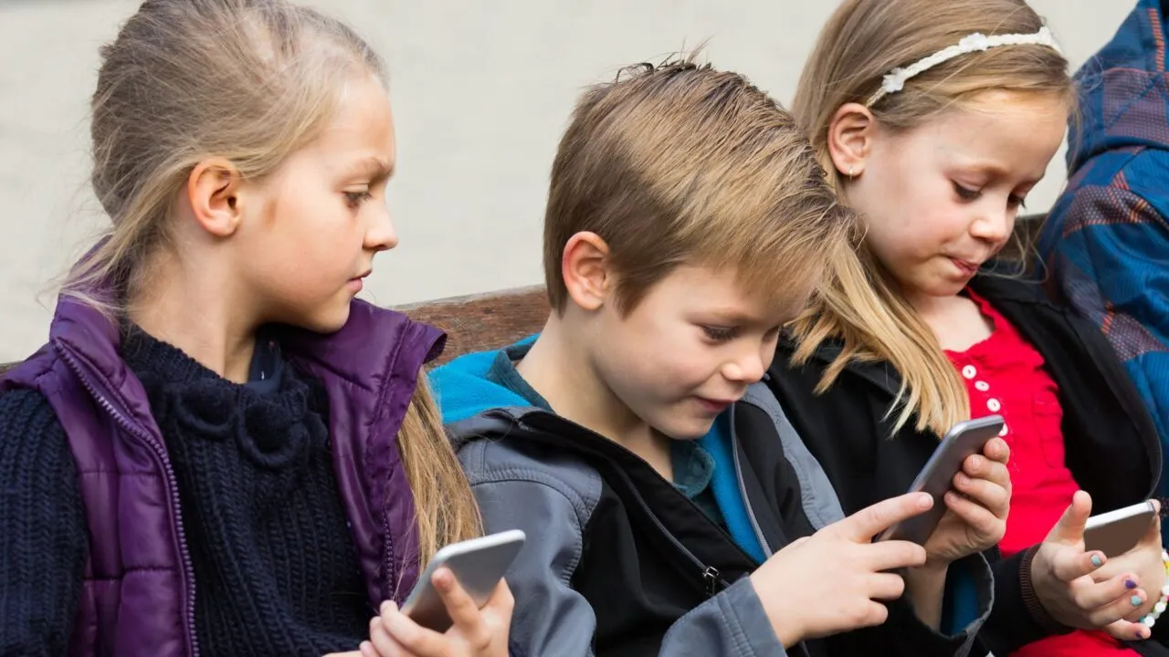 Tre børn sidder tæt på hinanden på en bænk med fokus på hver deres smartphone. De ser ud til at være optaget af deres skærme og interagerer ikke med hinanden.