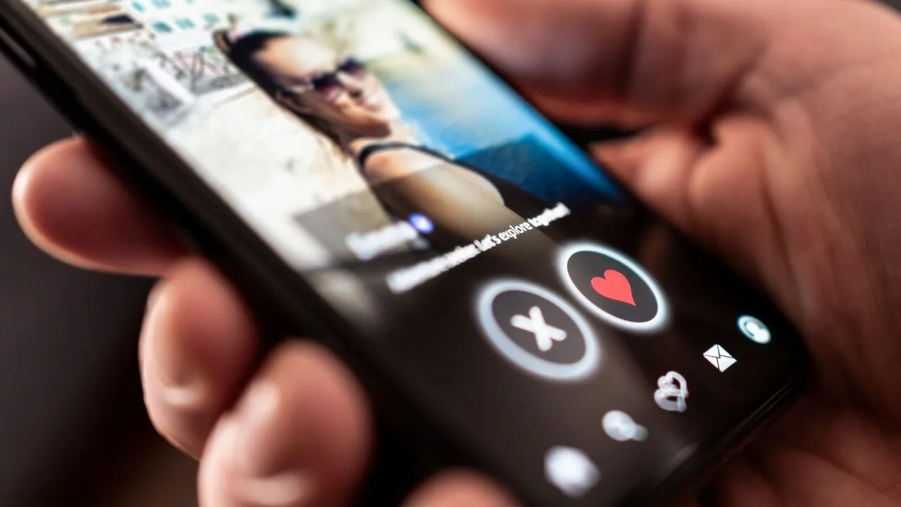 En person holder en smartphone, der viser en datingapp-profil med et sløret foto af en kvinde og synlige like- og dislike-knapper formet som et hjerte og et X.