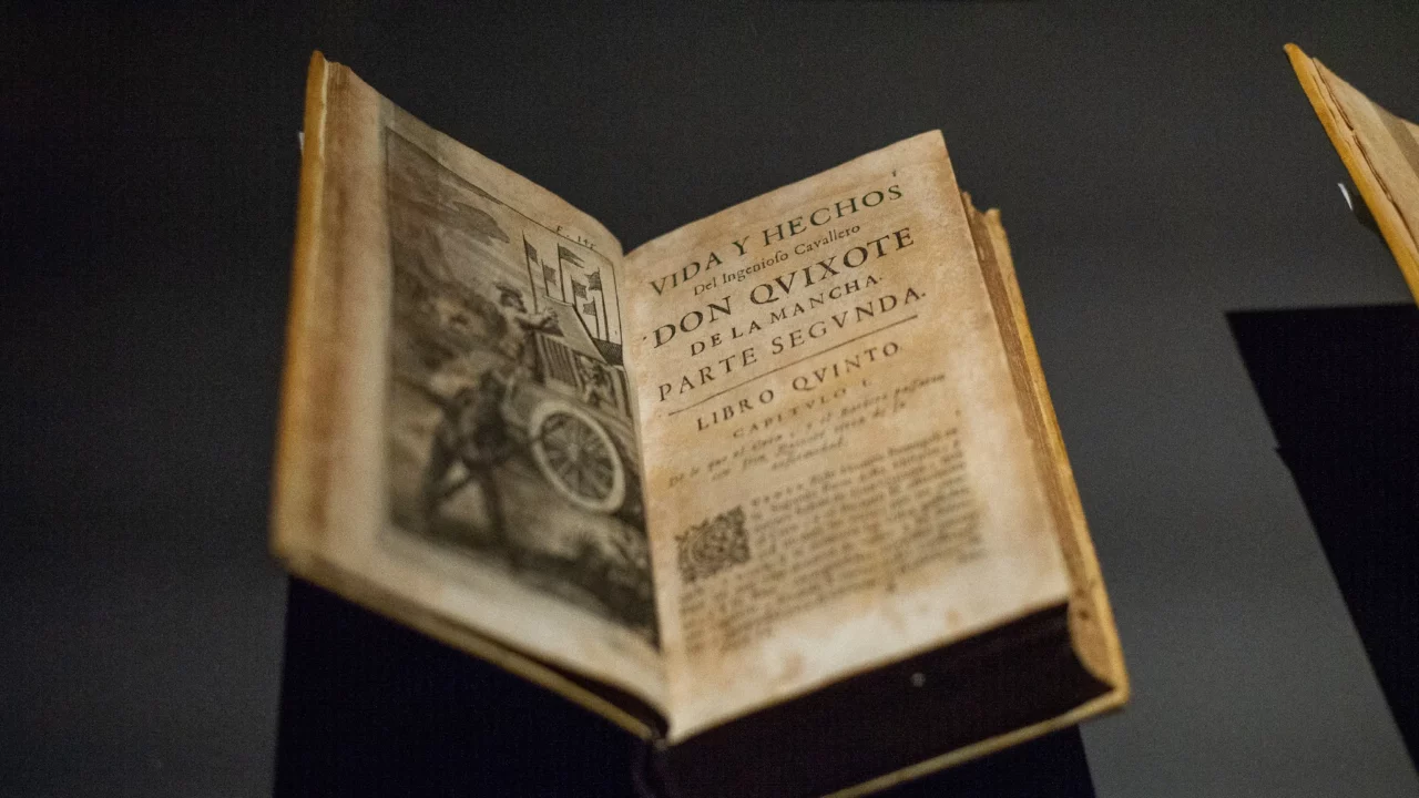 En gammel, opslået bog, der vises under et lys, med titelbladet og en illustration fra "Don Quijote de la Mancha, Parte Segunda". Siderne ser gamle og slidte ud.