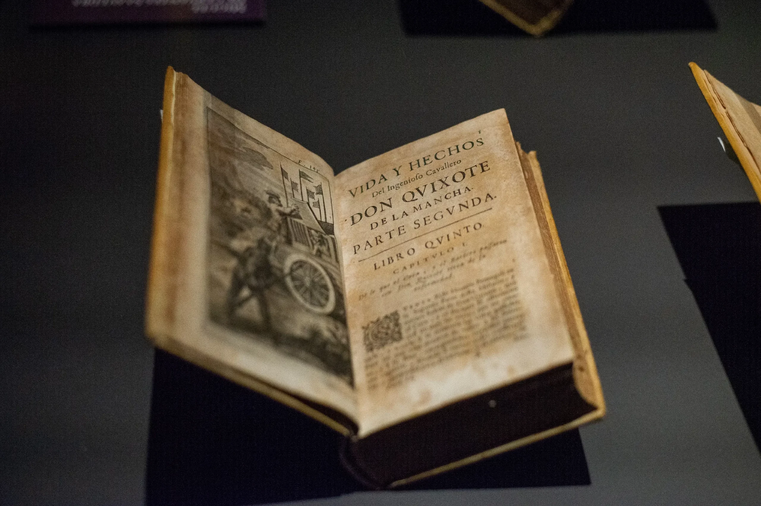 En gammel, opslået bog, der vises under et lys, med titelbladet og en illustration fra "Don Quijote de la Mancha, Parte Segunda". Siderne ser gamle og slidte ud.