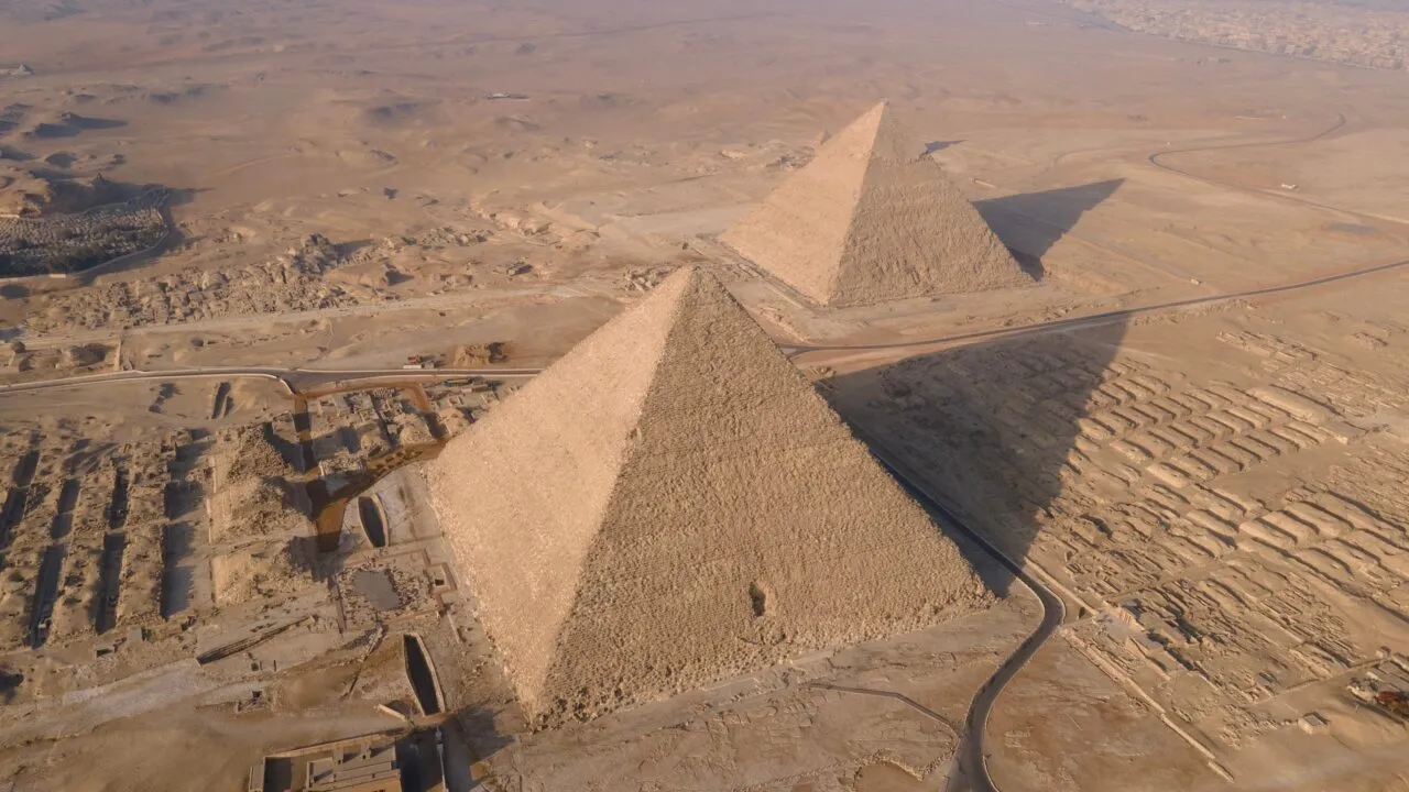 Luftfoto af pyramiderne i Giza i Egypten, omgivet af ørkensand med veje og gamle ruiner i nærheden, under en klar himmel.