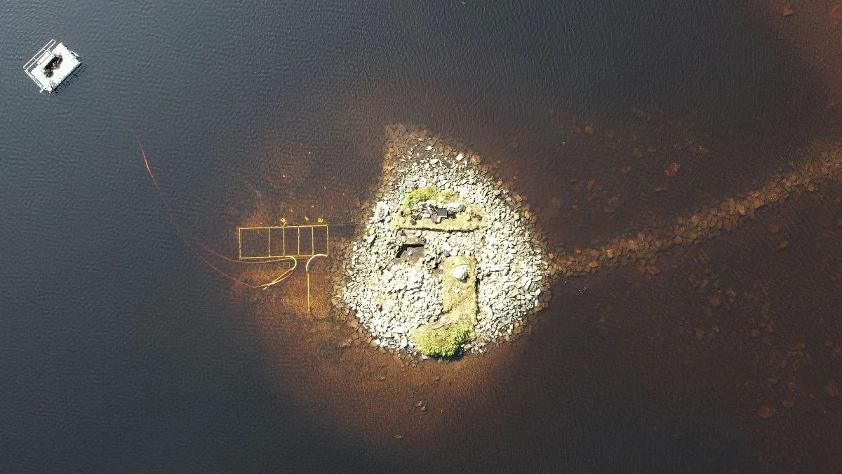 Luftfoto af en lille klippeø omgivet af mørkt vand med pletter af græs og ruinlignende strukturer. En svag, lige linje og en rektangulær ramme er synlig i vandet til venstre for øen.