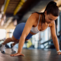 En kvinde i sportstøj laver en push-up på gulvet i et fitnesscenter, mens hun koncentrerer sig om sin træning. I baggrunden ses træningsudstyr og naturligt lys.
