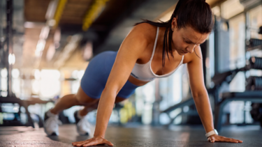 En kvinde i sportstøj laver en push-up på gulvet i et fitnesscenter, mens hun koncentrerer sig om sin træning. I baggrunden ses træningsudstyr og naturligt lys.