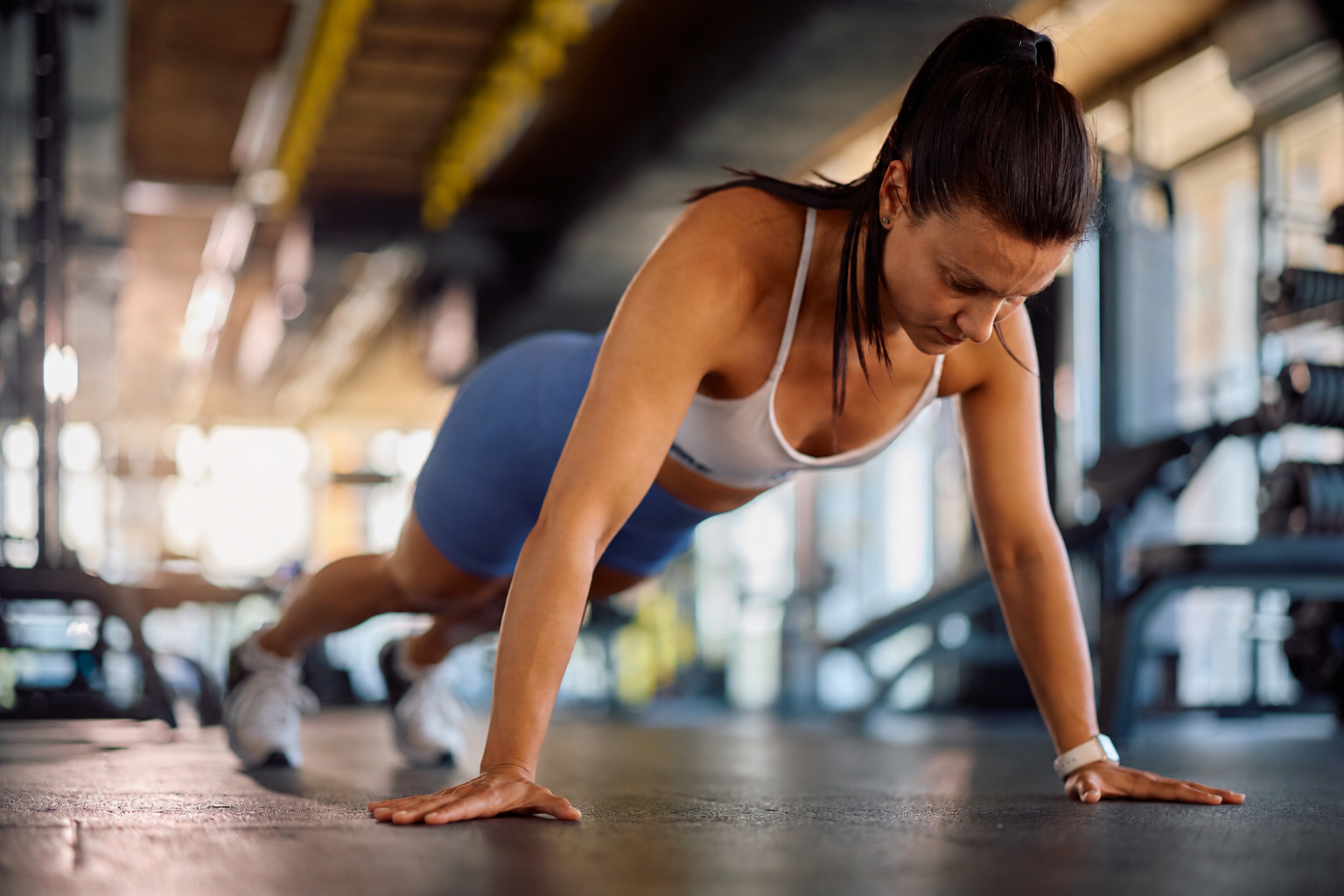 En kvinde i sportstøj laver en push-up på gulvet i et fitnesscenter, mens hun koncentrerer sig om sin træning. I baggrunden ses træningsudstyr og naturligt lys.
