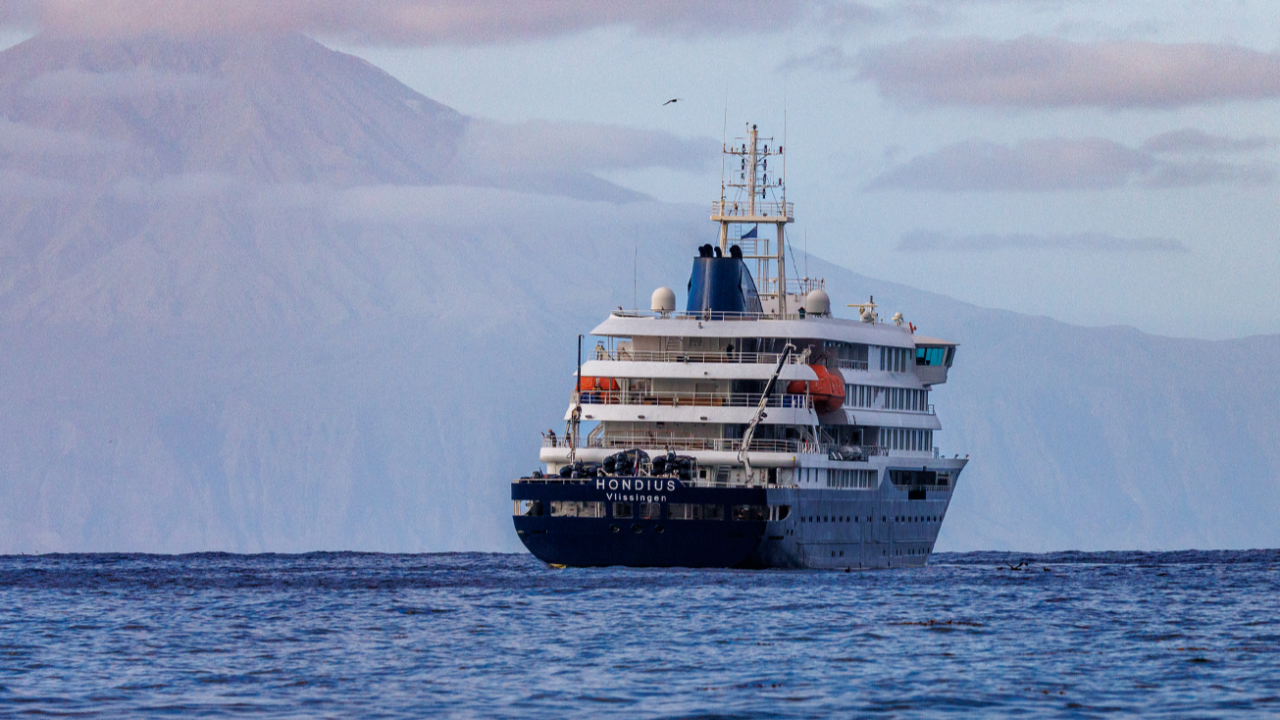 Et stort hvidt og blåt krydstogtskib ved navn Hondius sejler på roligt havvand med et bjergrigt landskab og en overskyet himmel i baggrunden.