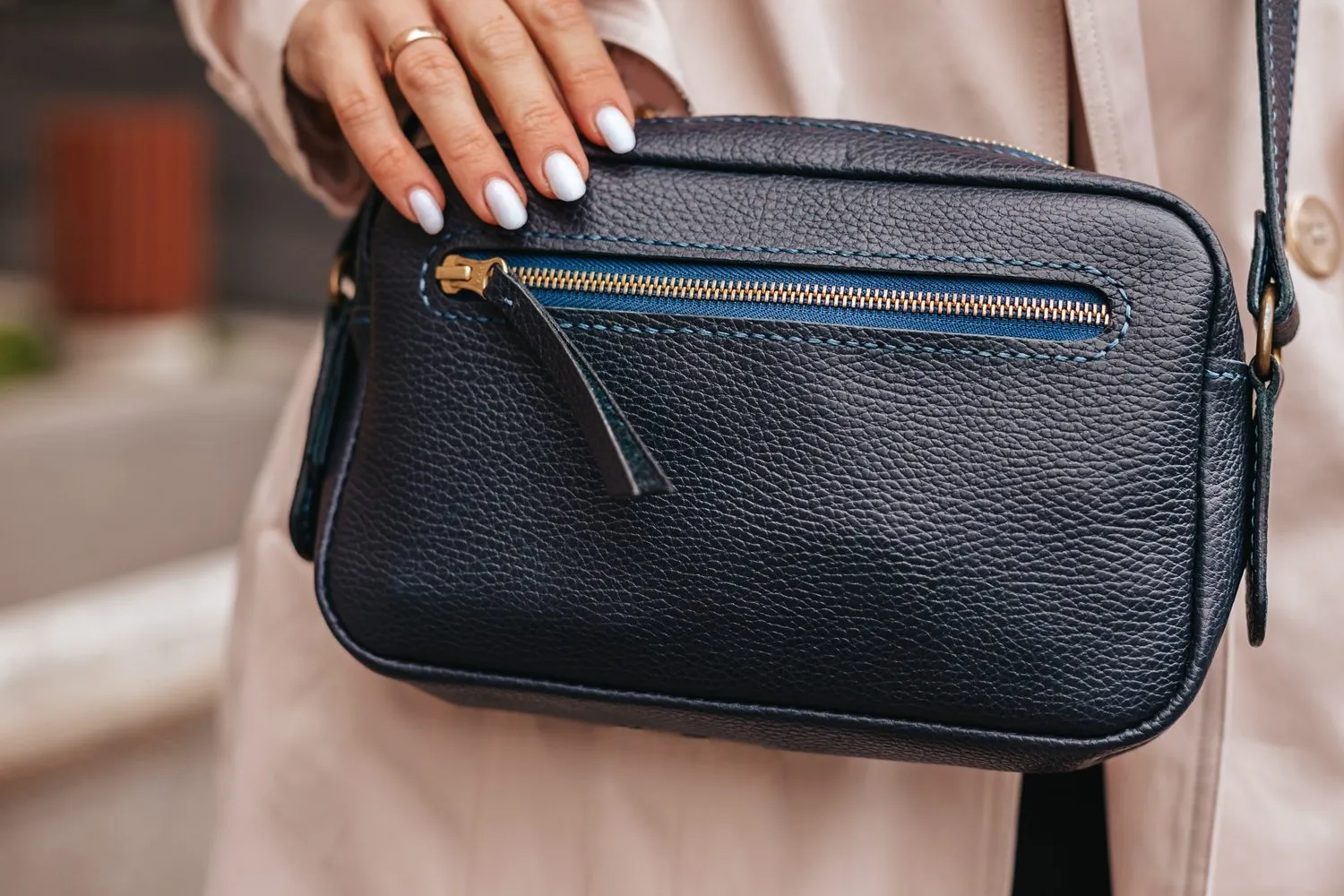 En person med hvidlakerede negle og en guldring holder en crossbody-taske i sort struktureret læder med en lynlåslomme foran og er iført en beige frakke.