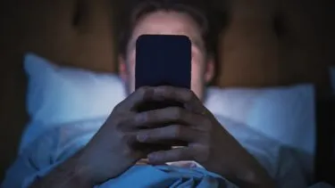 En person ligger i sengen om natten og holder og kigger på en smartphone. Ansigtet er svagt oplyst af telefonens skærm, mens resten af rummet er mørkt.