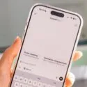 En hånd holder en smartphone, der viser ChatGPT-appens grænseflade med muligheder som Create a painting og Summarize a long document synlige på skærmen. I baggrunden ses en sløret computerskærm.