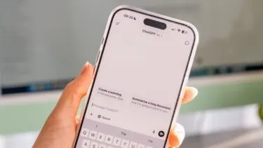 En hånd holder en smartphone, der viser ChatGPT-appens grænseflade med muligheder som Create a painting og Summarize a long document synlige på skærmen. I baggrunden ses en sløret computerskærm.