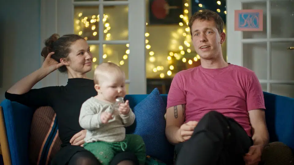 En kvinde, en mand og en baby sidder sammen på en blå sofa i et hyggeligt rum med lyskæder i baggrunden. Kvinden smiler til manden, mens babyen holder en ske og kigger ned.