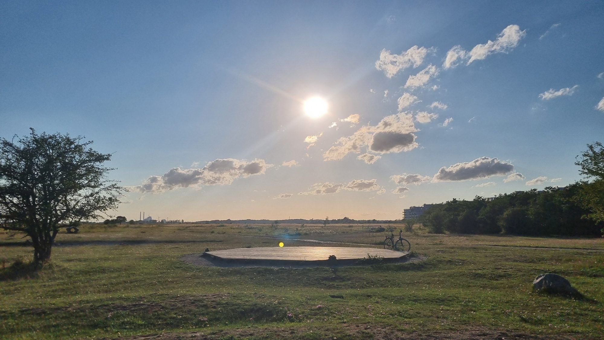 En rund betonplatform ligger på en græsplæne under en klar himmel med spredte skyer. En cykel står lænet op ad platformen, med træer og skygger i baggrunden. Solen skinner over horisonten.