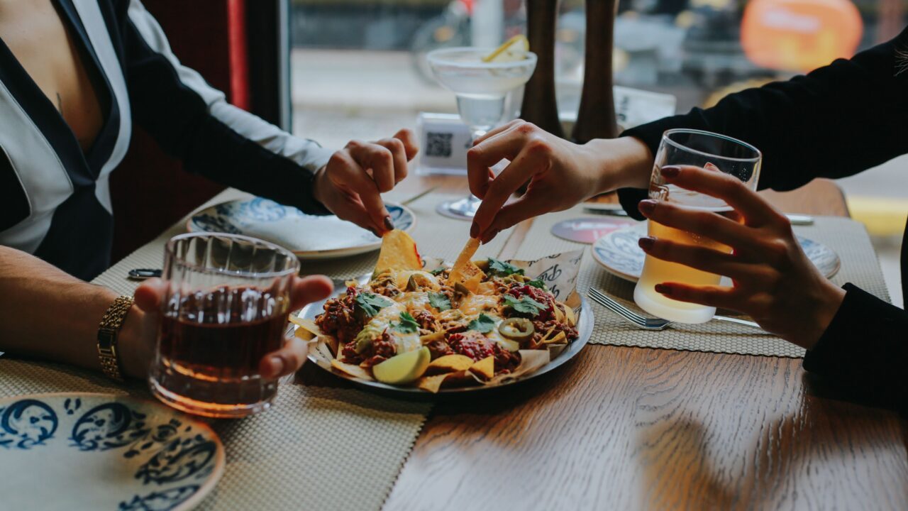 To personer sidder ved et bord på en restaurant, hvor de deler en tallerken med nachos med masser af tilbehør og holder drinks i hånden. Deres hænder rækker ud efter maden, og bordet er dækket med tallerkener, drinks og krydderier.