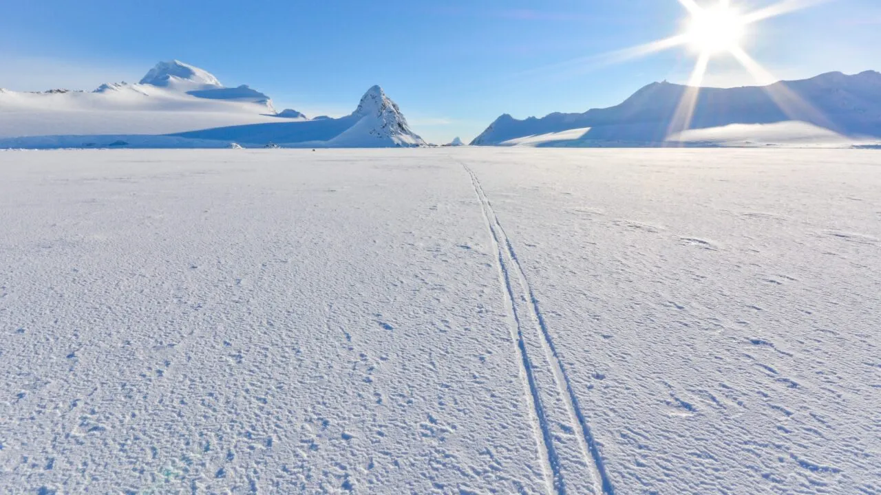 En strålende sol skinner over et vidtstrakt, snedækket landskab med skispor, der snor sig ud i det fjerne, omgivet af bjerge under en klar, blå himmel.