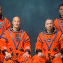 Fire astronauter i knaldorange NASA-rumdragter poserer mod en mørkeblå baggrund; to sidder og holder hjelme, og to står bag dem og holder også hjelme.