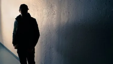 En person i kasket og jakke står i skygge mod en tekstureret væg, hvor lyset oplyser en del af baggrunden og skaber en dramatisk og mystisk atmosfære.