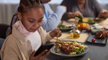 En ung pige med fletninger kigger på sin smartphone, mens hun spiser sammen med andre ved et bord. Hendes tallerken indeholder pasta, salat og kød, og der står mere mad og drikkevarer på bordet.