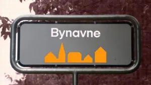 Et gadeskilt med ordet Bynavne over orange silhuetter af bygninger, monteret på en metalramme, med træer og blade sløret i baggrunden.