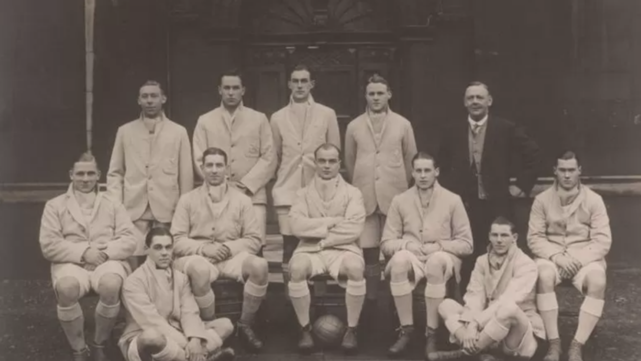 Et gammelt sort-hvidt foto af elleve mænd i ens lyse sportsdragter og en mand i jakkesæt, der poserer som et fodboldhold foran en stor buet døråbning. Der ligger en fodbold på jorden foran dem.