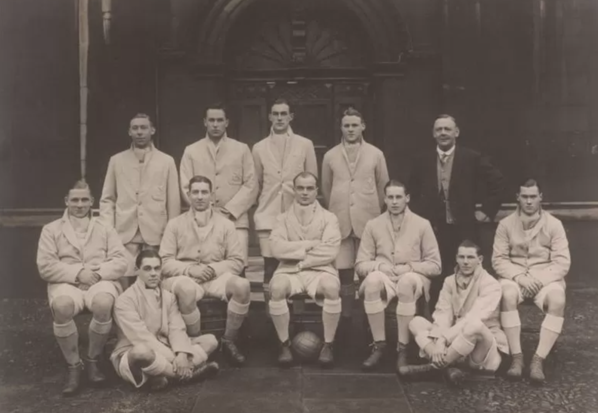 Et gammelt sort-hvidt foto af elleve mænd i ens lyse sportsdragter og en mand i jakkesæt, der poserer som et fodboldhold foran en stor buet døråbning. Der ligger en fodbold på jorden foran dem.