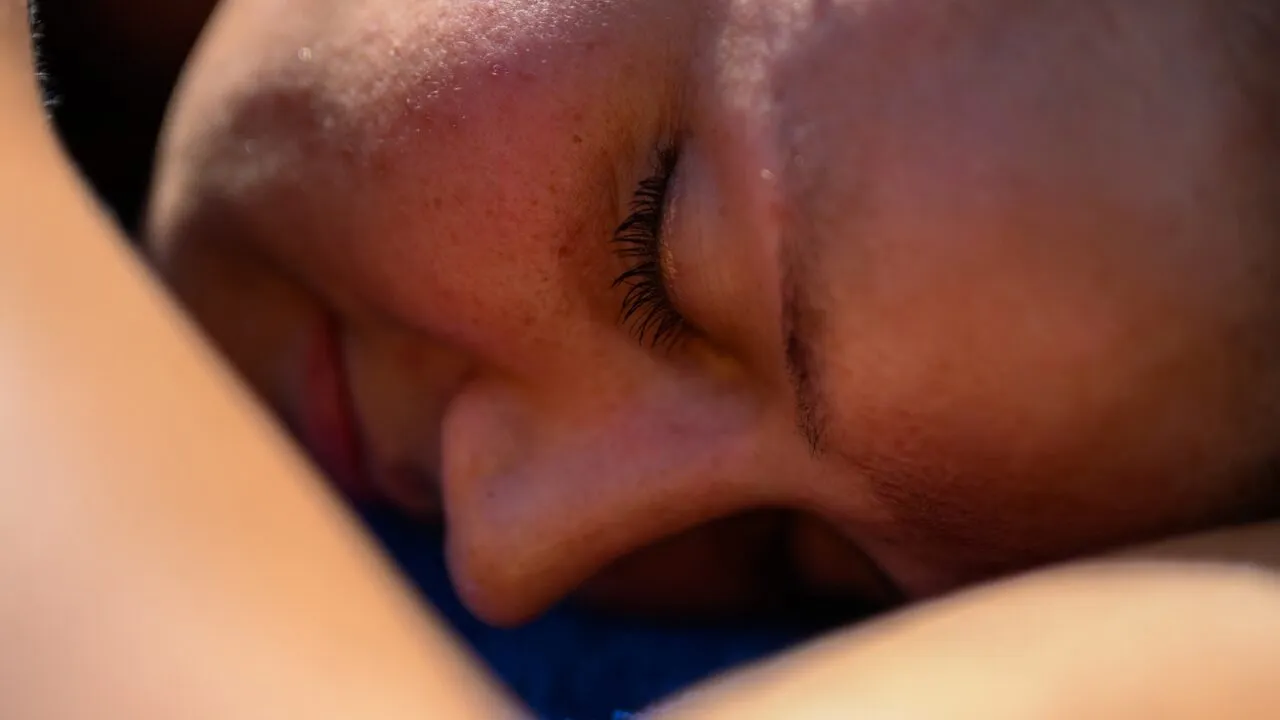 Nærbillede af en persons ansigt, der hviler på en overflade, med lukkede øjne, hvor det naturlige lys fremhæver øjenvipper, næse og kind. Billedet udstråler en følelse af afslapning og ro.