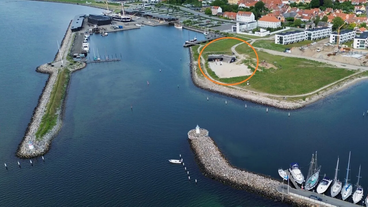 Luftfoto af en kystby med en lystbådehavn, både, der ligger fortøjet langs molerne, og et sandområde, der er markeret med en orange cirkel, tæt på et græsareal og bygninger i baggrunden.