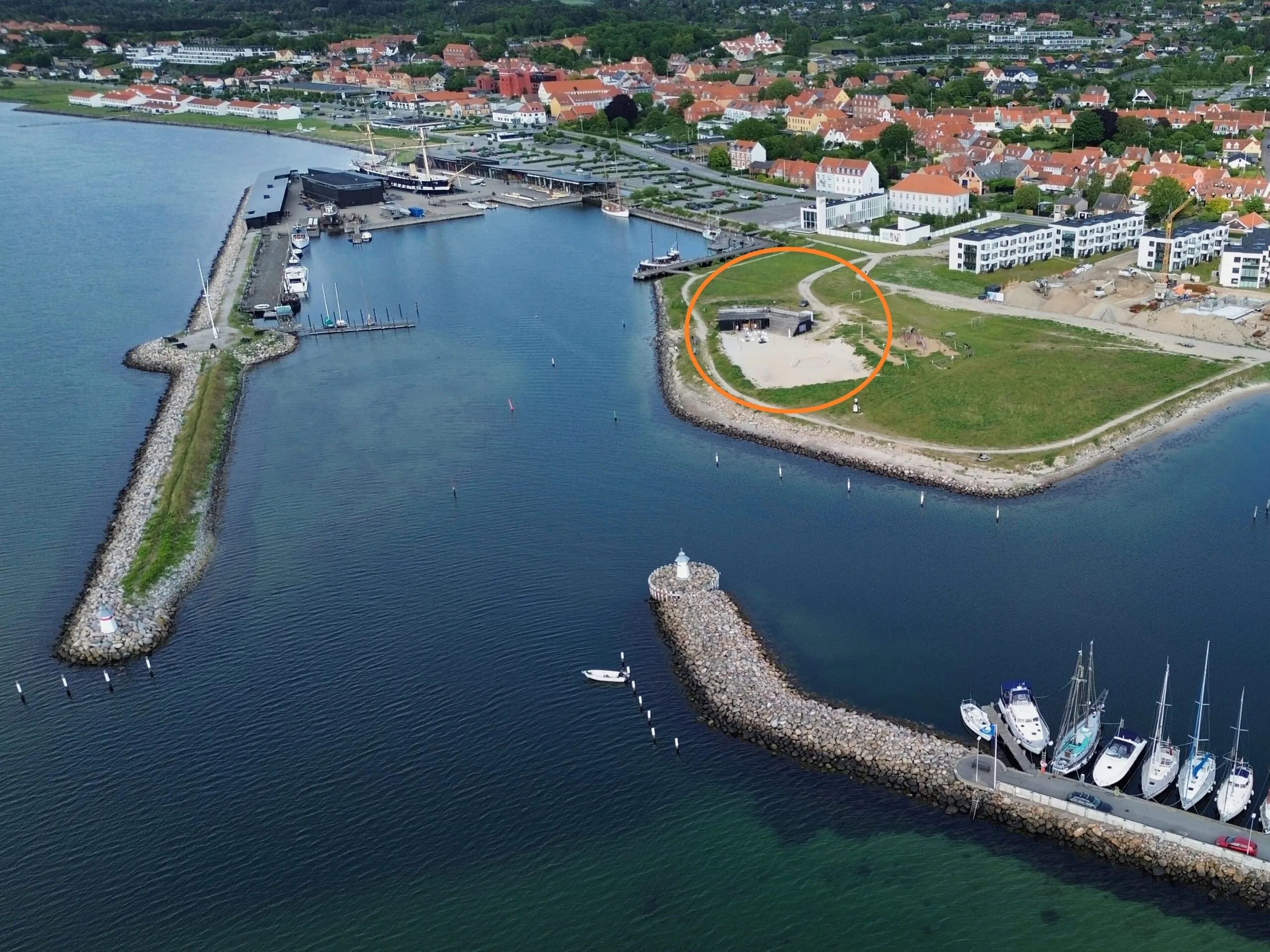 Luftfoto af en kystby med en lystbådehavn, både, der ligger fortøjet langs molerne, og et sandområde, der er markeret med en orange cirkel, tæt på et græsareal og bygninger i baggrunden.